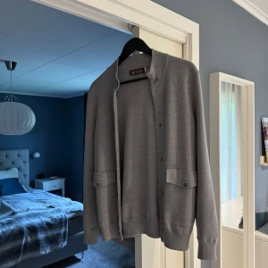 Grå kofta från Morris - Stilren grå kofta från Morris med knappar framtill och två fickor. Tröjan har lång ärm och ribbade muddar vid ärmslut och nederkant. Perfekt för lager-på-lager och enkel att matcha.