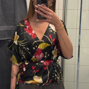 Svart blommig omlottblus från Vero Moda - Säljer en svart omlottblus från Vero Moda med färgglatt blommönster i rött, gult och grönt. Blusen har v-ringning, korta ärmar och knytning i midjan för en snygg passform. Perfekt för att liva upp garderoben!