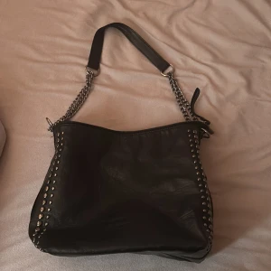 Svart axelväska med nitar och kedjedetaljer - Säljer en svart axelväska med coola silverfärgade nitar längs sidorna och kedjor som axelrem. Väskan har en modern look och är perfekt för att lyfta vilken outfit som helst. Rymmer det mesta du behöver till vardags.