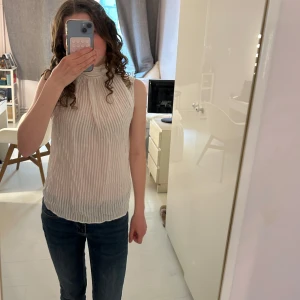Vit ärmlös blus med rosett i ryggen - Säljer en vit ärmlös blus med diskreta ränder och en stor rosett i ryggen. Blusen har hög hals och är tillverkad i ett lätt, luftigt material som passar perfekt till varmare dagar.