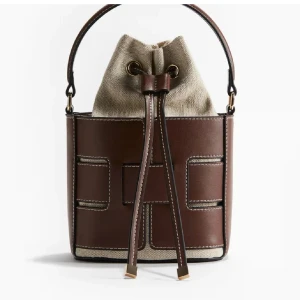 Hm väska  - Trendig bucket bag i brunt skinn och beige canvas med snygga detaljer. Väskan har snörning upptill och ett stadigt handtag. Perfekt accessoar för att lyfta din outfit. Väskan är slutsåld !! Prislapp finns kvar!