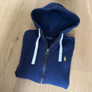Marinblå hoodie från Polo Ralph Lauren - Säljer en marinblå hoodie från Polo Ralph Lauren med dragkedja och huva. Tröjan har vit dragsko och den klassiska gula broderade loggan på bröstet. Tillverkad i mjuk bomull, perfekt för en avslappnad stil.