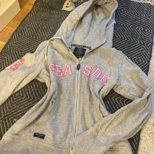 Grå ziphoodie från Seaside - Grå hoodie med dragkedja från Seaside. Har huva med dragsko och rosa text på bröstet samt ärmen. Perfekt för en avslappnad stil och enkel att matcha med jeans eller leggings.