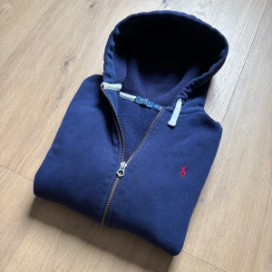 Marinblå hoodie från Polo Ralph Lauren - Säljer en marinblå hoodie från Polo Ralph Lauren med dragkedja och vit snörning i huvan. Tröjan har det klassiska röda Polo-logot broderat på bröstet och är tillverkad i mjuk bomull. Perfekt för en avslappnad och stilren look.
