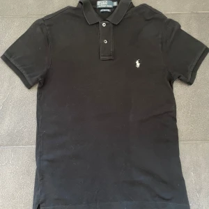 Ralph Lauren Pike  - Klassisk svart pikétröja från Polo Ralph Lauren med vit broderad logga på bröstet. Tröjan har korta ärmar, krage och knäppning med två knappar. Slim fit-modell i stretchigt material.           Storlek: M.                                                                        Skick: Mycket bra🤗