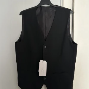 Svart kostymväst från Selected Homme - Stilren svart kostymväst från Selected Homme med klassisk v-ringning och knäppning framtill. Västen har släta fickor och är helfodrad med en elegant finish. Perfekt för att lyfta din outfit med en tidlös touch.