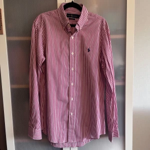 Röd- och vitrandig skjorta från Ralph Lauren - Säljer en klassisk röd- och vitrandig skjorta från Ralph Lauren med broderad logga på bröstet. Skjortan har lång ärm, knappar framtill och är i bomull. Custom fit-modell som passar perfekt till många tillfällen.