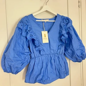 Blå blus med volang och broderade detaljer - Säljer en blå blus från Pernilla Wahlgren med v-ringning, volanger och broderade detaljer upptill. Blusen har trekvartsärmar med puff och resår i midjan för en fin siluett. Perfekt för vår och sommar!