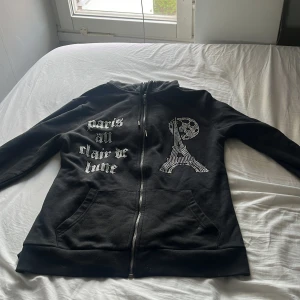 Svart hoodie med Paris-tryck och strassdetaljer - Svart hoodie med dragkedja och huva, dekorerad med texten 'Paris au clair de lune' och en Eiffeltorn-design i strass på framsidan. Har fickor framtill och snörning i huvan. Perfekt för en avslappnad stil.