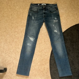 Replay Jeans - Säljer ett par blå Replay Anbass jeans med snygga slitningar och raka ben. Jeansen har klassisk femficksdesign, dragkedja och Replay-logga på bakfickan. Perfekta för dig som gillar en modern och avslappnad stil. Riktigt bra skick. Inga defekter eller så. Nypris runt 1699kr mitt pris 500kr. Kontakta mig om du har frågor✅