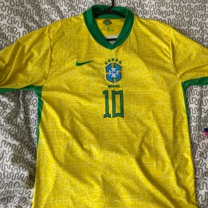 Brasilien fotbollströja nummer 10 - Säljer en gul Brasilien fotbollströja från Nike med gröna detaljer och nummer 10 på bröstet. Tröjan har korta ärmar och ett subtilt mönster över hela framsidan. Perfekt för dig som älskar fotboll och vill visa ditt stöd för Brasilien. Neymar JR 10