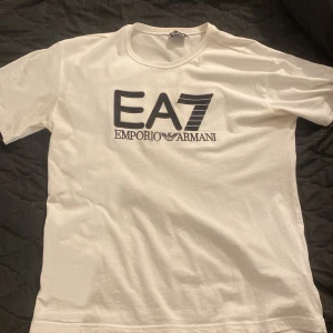 Vit t-shirt från Emporio Armani EA7 - (ANDVÄNT 1 GÅNG)                                       Säljer en vit t-shirt från Emporio Armani EA7. med stor svart logga på bröstet.  Perfekt för dig som gillar stilrena märkesplagg. 100% äkta köpt på NK