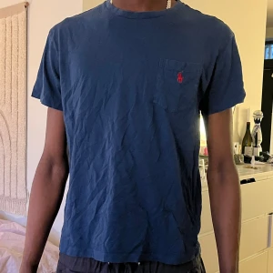 Blå t-shirt från Ralph Lauren - Mörkblå t-shirt från Ralph Lauren med liten röd logga broderad på bröstfickan. Klassisk rund halsringning och korta ärmar. Perfekt basic-plagg med enkel och stilren design.