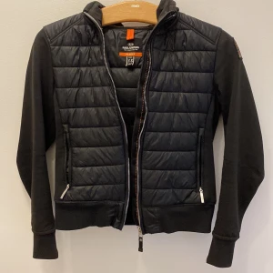 Svart hybridjacka från Parajumpers - Svart jacka från Parajumpers med quiltad front och rygg samt mjuka ärmar och huva. Jackan har dragkedja framtill och två fickor med dragkedja. Perfekt för lager-på-lager och har en sportig look.