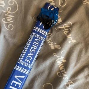 Blått bälte från Versace med Medusa-spänne - Säljer ett blått bälte från Versace med tydlig logga längs bandet och ett blått Medusa-huvud som spänne. Bältet har en modern och iögonfallande design, perfekt för dig som vill sticka ut.