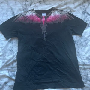 Svart t-shirt med vingmönster från Marcelo Burlon - Säljer en svart t-shirt från Marcelo Burlon med ikoniskt vingmönster i rosa och grått över axlar och halsringning. T-shirten är i mjuk bomull och har en normal passform med korta ärmar. 