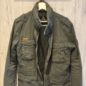 Grön field-jacket från Superdry - Säljer en grön militärinspirerad jacka från Superdry Rookie Edition. Jackan har flera fickor framtill, axelklaffar och dragkedja med knappslå. Perfekt för dig som gillar en stilren och funktionell look.