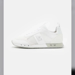 Vita sneakers från Emporio Armani EA7 - Snygga vita sneakers från Emporio Armani EA7 med meshpaneler och detaljer i syntet. Skorna har snörning och en platt sula med räfflad undersida. Perfekta för en stilren och sportig look.