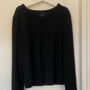 Svart kofta från Gina Tricot - En klassisk svart kofta från Gina Tricot med rund halsringning och en knapp framtill. Koftan har lång ärm och är tillverkad i ett mjukt, tunt material som passar perfekt för lager-på-lager. Säljer för den inte kommer till användning 