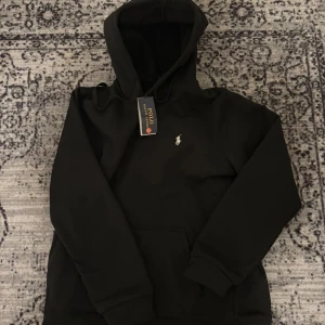 Svart hoodie från Polo Ralph Lauren - Säljer en svart hoodie från Polo Ralph Lauren med klassisk broderad logga på bröstet. Tröjan har huva med dragsko och en stor magficka framtill. Perfekt för en stilren och avslappnad look.