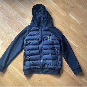 Svart cardigan jacka med huva från Garde Grisch - Säljer en svart pufferjacka från Garde Grisch i storlek XS. Jackan har huva, dragkedja framtill och vadderade paneler på bröst och rygg. Logga på bröstet och ärmen. Perfekt för kyliga dagar.