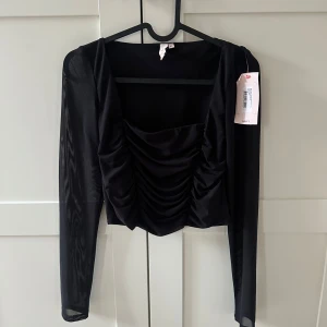 Svart mesh topp med rynkade detaljer - Trendig svart topp från Nelly med långa meshärmar och rynkade detaljer framtill. Fyrkantig ringning och figurnära passform ger en snygg siluett. Perfekt för att lyfta din outfit. Aldrig använd!