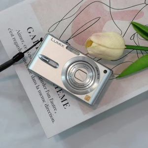 Lumix Fx77 - Ingen väska syns på bilden. Bilden visar en silverfärgad digitalkamera från Lumix placerad på ett papper med en tulpan bredvid.