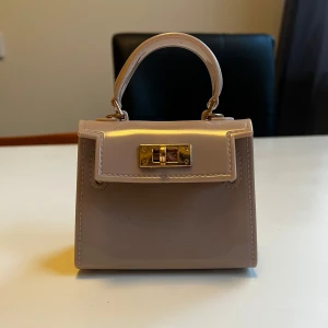 Beige mini handväska med gulddetalj - Söt beige mini handväska med stilren design och guldfärgat lås framtill. Väskan har ett kort handtag och är tillverkad i glansigt syntetmaterial. Perfekt accessoar för att lyfta din outfit.