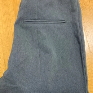 Grå kostymbyxor från H&M - Snygga grå kostymbyxor från H&M med klassisk rak passform. Byxorna har pressveck och en diskret bakficka utan synlig knapp. Perfekta för en stilren look.