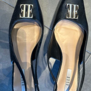 Svarta klackskor med metallogo från ELLE - Säljer ett par svarta klackskor från ELLE med öppen häl och spetsig tå. Skorna har en elegant metallogo framtill och en låg klack, perfekt för att lyfta din outfit.