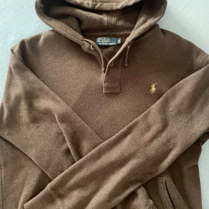 Brun hoodie från Polo Ralph Lauren - Säljer en brun hoodie från Polo Ralph Lauren med klassisk broderad logga på bröstet. Tröjan har huva med dragsko, knappar vid halsen och är tillverkad i 100% bomull. Perfekt för en avslappnad stil.