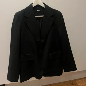 Svart kavaj från Gina Tricot - Klassisk svart kavaj från Gina Tricot med två fickor framtill och knäppning med en knapp. Perfekt för en stilren look och passar till många olika outfits. Använd 1 gång (ordinarie pris 599kr)