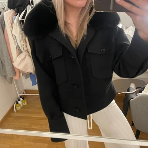 Svart jacka med fuskpäls-krage - Säljer en svart jacka med stor fuskpäls-krage och stora fickor framtill. Jackan har knappar hela vägen och en boxig, croppad passform. Perfekt för dig som vill ha en stilren och trendig look.