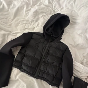 Svart pufferjacka med huva - Säljer en svart pufferjacka med huva och dragkedja framtill. Jackan har quiltade partier på kroppen och släta, långa ärmar. Perfekt för dig som vill ha en cool och sportig look.