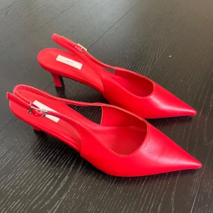 Röda pumps med öppen häl från Nelly - Säljer ett par klarröda pumps från Nelly med spetsig tå och öppen häl. Skorna har en smal klack och justerbart spänne vid ankeln. 