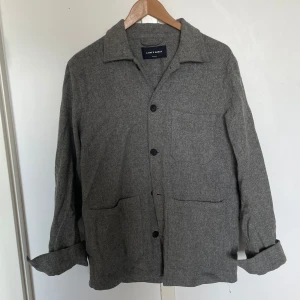Grå overshirt från A Day's March - Stilren grå overshirt från A Day's March med klassisk krage och tre framfickor. Jackan har knäppning framtill och är tillverkad i ett mjukt ullmaterial. Perfekt lager för kyligare dagar.