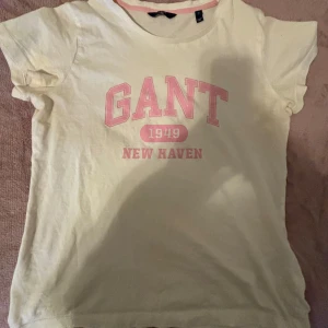 Gul t-shirt från GANT med tryck - Snygg gul t-shirt från GANT med rosa tryck framtill där det står 'GANT 1949 NEW HAVEN'. Klassisk rund halsringning och korta ärmar. Perfekt för en avslappnad stil och enkel att matcha med jeans eller shorts. Tyvär har den dok någon svett fläck från tidigare ägaren, Men själv har jag inte använt den så mycket men när jag gjort det så enligt mig märks det inte så mycket. Jag har tvättat den några gånger i tvätten men har inte fukuserat på att få bort det så vet inte riktigt om det går bort.💗💖💕