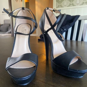 Svarta platåklackar från Bebo - Säljer ett par svarta höga klackar från Bebo med platåsula och öppen tå. Skorna har smala remmar som korsas över foten och runt vristen, samt spänne vid ankeln. Perfekta för dig som gillar att sticka ut med höga klackar.