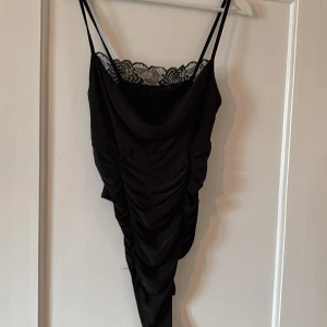 Svart body med spets från Boohoo - Supersnygg svart body från Boohoo med smala axelband och spetsdetalj upptill. Klänningen har omlott-effekt och rynkade detaljer framtill som ger en cool look. Perfekt för dig som vill sticka ut lite extra!
