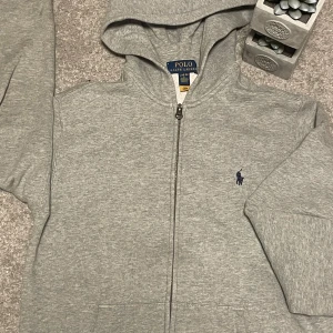 Grå Ralph lauren Zipp hoodie - Säljer en klassisk grå hoodie från Polo Ralph Lauren med dragkedja och huva. Tröjan har broderad logga på bröstet och två fickor framtill. Perfekt för en avslappnad stil.