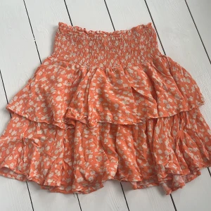 Volangkjol från Samsa Collection gekås - Supersöt kort volangkjol från Samsa Collection i orange med vita blommiga mönster. Kjolen har smock i midjan för en bekväm passform och flera lager volanger som ger en riktigt flowy känsla. 