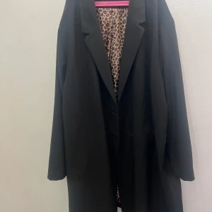 Svart kappa från Ellos med leopardfoder - Svart kappa från Ellos i klassisk rak modell med snyggt leopardmönstrat foder. Jackan har långa ärmar, två fickor framtill och stilren krage. Perfekt för dig som vill ha en tidlös look med en cool twist på insidan.