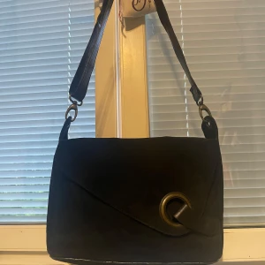 Svart väska - Elegant axelväska i svart mocka från Vera Pelle – tillverkad i Italien. Modern design med asymmetrisk framsida och ett stilrent lock som pryds av en stor metallring. Tillverkad i mjuk mocka med struktur och djup färgton för ett sofistikerat intryck. Bred axelrem för komfortabel passform och en minimalistisk silhuett som passar både vardag och kväll. Diskret Vera Pelle-logotyp.