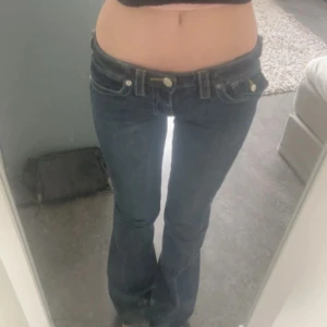 True Religion Joey Low Rise Flare jeans - Säljer ett par blå True Religion Joey jeans med låg midja och utsvängda ben. Klassiska kontrastsömmar, stora fickor bak med vita knappar och broderade detaljer. Jeansen är i denim och har en snygg flare fit som ger en retro vibe.