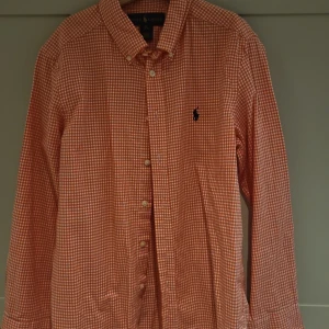 Rutig orange skjorta från Ralph Lauren - Snygg orange och vit rutig skjorta från Ralph Lauren med klassisk krage och knäppning framtill. Skjortan har lång ärm och ikonisk broderad logga på bröstet. Perfekt för dig som vill ha en stilren och tidlös look.