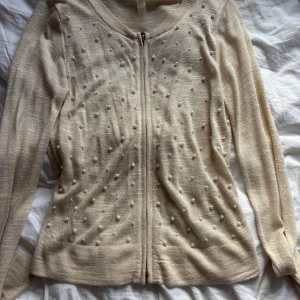 Beige kofta med pärlor från Guess - Supersöt beige kofta från Guess med dragkedja framtill och pärldetaljer över hela framsidan. Långärmad och perfekt för lager-på-lager. Snygg och feminin look som lyfter vilken outfit som helst.