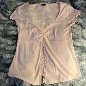 Ljusrosa v-ringad topp med spets - Supersöt ljusrosa topp med v-ringning och korta ärmar. Toppen har en snygg rynkdetalj framtill och spetskant längs urringningen och mittsömmen. Perfekt för dig som gillar romantiska detaljer och mjuka färger.