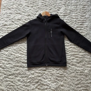 Svart hoodiejacka från Sail Racing - Svart hoodiejacka från Sail Racing med dragkedja framtill och bröstficka med logga. Jackan har huva med broderad detalj och är tillverkad i mjukt bomullsmaterial. Perfekt för dig som gillar stilren och sportig look.