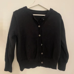 Svart stickad kofta med knappar - En klassisk svart stickad kofta med rund hals och knäppning framtill. Koftan har långa ärmar och ribbade muddar vid ärmslut och nederkant. Perfekt att slänga över en t-shirt eller klänning. 