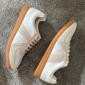Grisch sneakers beige/vit - Grisch sneakers i vitt och beige skinn och mocka med klassisk gummisula i brunt. Snygg retrostil med rund tå, platt sula och snörning. Perfekta för dig som gillar minimalistisk och clean design med lyxig känsla. Pris kan diskuteras